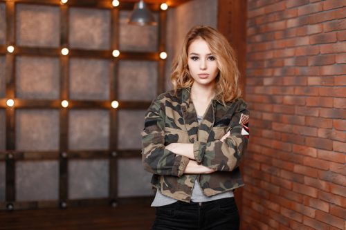 A young, stylish woman leaning against a brick wall, wearing a camouflage jacket