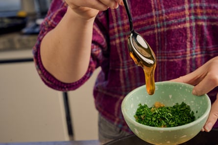 A hand drizzling a tablespoon of honey into a small bowl containing chopped parsley.