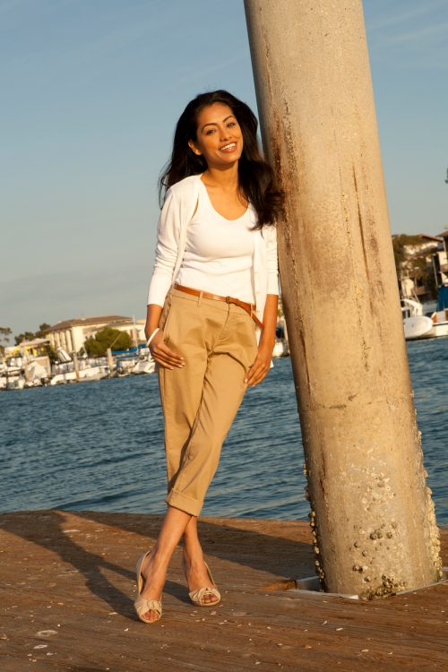 An attractive woman leaning against a post on a pier, wearing an off-white top and camel-colored capri pants