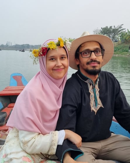 A woman in a headscarf and wearing a flower wreath on her head, sits happily next to a man in glasses and a brimmed hat, on a rowboat on a river.