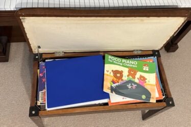 A piano stool, opened to reveal a pile of piano music books including one book illustrated with three happy bears and titled ‘Poco Piano for Young Children’.