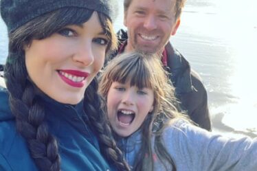 A woman in a beanie, with husband and  primary-school aged daughter pose happily for a selfie photo during an camping trip.