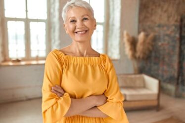 A mature woman with short white hair wearing a yellow-orange dress is standing in her living room with her arms crossed posing for a picture.