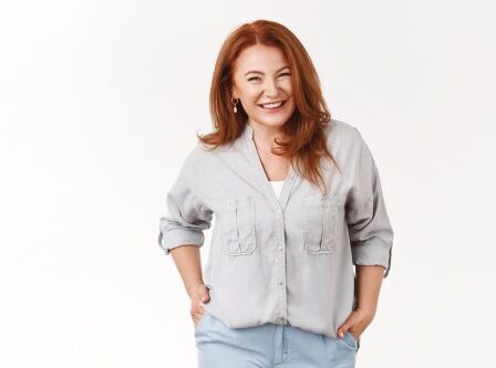 A cheerful woman with red hair wearing a light gray button-down shirt poses against a white background