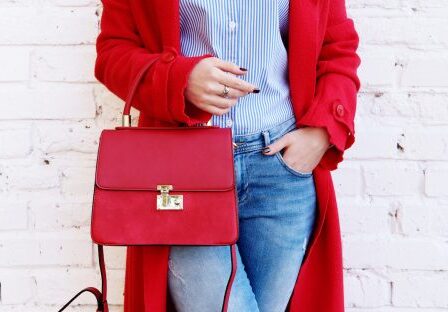 Closeup of red small bag in hand of woman. Fall spring fashion outfit red coat and trendy blue jeans
