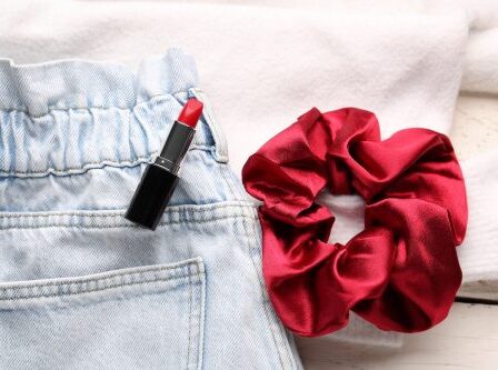 Close up of elastic-waist light-wash jeans, a red scrunchie, and red lipstick against a white background