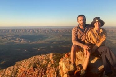 Tess and Piers at Mount Sonder, Northern Territory in 2022. They are seated on a rock lit up by the golden light of a setting sun shining from the left of the photo, while behind them can be seen an expansive green valley with a line of mountainous terrain near the horizon.