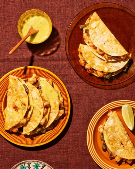 Tacos gobernador, or cheesy prawn tacos, piled two to three on a plate alongside a bowl of guacamole falso and a lime wedge. The plates are variously made of ochre-coloured glass or ceramic.