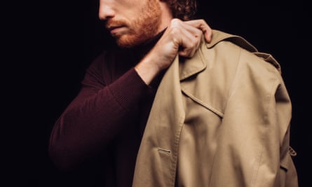 Studio photo of a ginger man model holding a trench coat on his shoulder.