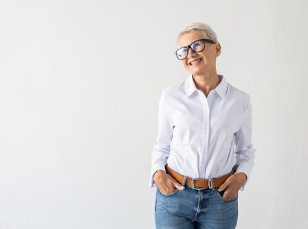 A mature woman with short white hair is wearing a white button-down shirt with her hands in her jeans pockets against a white background.