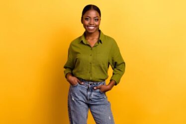 A young woman wearing an olive green shirt and jeans stands against a yellow background.