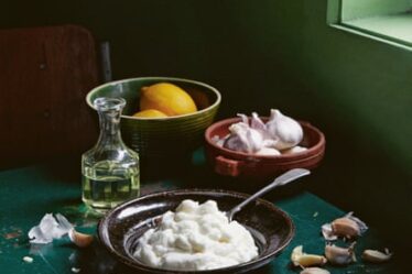Three earth-toned bowls set on a dark green table; they are (from top to bottom) holding lemons, garlic, and fluffy toum. The table is also scattered with garlic cloves. Next to the bowls is a glass bottle of oil.