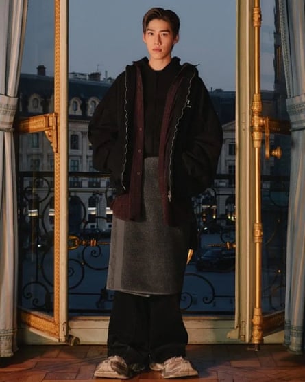 A male model poses in front of French doors wearing a towel over trousers with a large jacket on top.