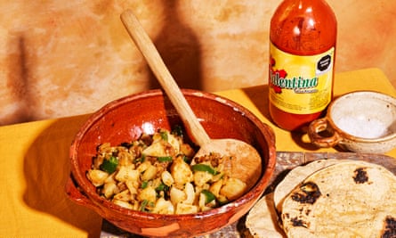 A photo of tacos de papas con cebolla, with the potato, chili and onion mixture in an earthen red bowl next to a stack of slightly charred corn tortillas. Behind the dishes is a bottle of Valentina’s hot sauce.