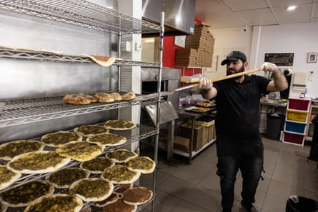 A male baker holds a long paddle, sliding just-baked manoush (Lebanese pizza) on to a bakery shelf.