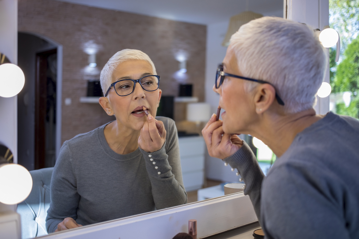 Portrait of smiling senior woman with short grey hair looking at herself in the mirror and putting lipstick on.