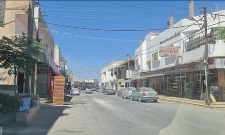 A dusty street scene with shops with signs in Arabic and lots of overhead wires and cars parked on the side of the road