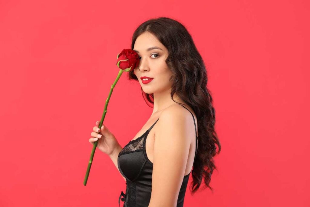 Elegant woman in black lingerie with a red rose on a pink background.