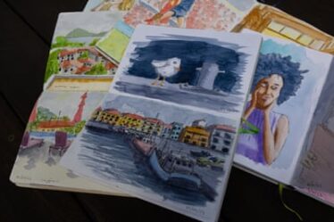 A sketchbook sits on top of a pile of other sketchbooks, with a seagull painted on one page and boards in a harbour on another.