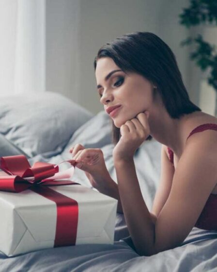 Contemplative woman in red lace lingerie admiring a wrapped gift on the bed.