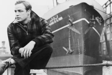 Marlon Brando pictured in black and white, in On the Waterfront, 1954, sitting on railings in front of a large ship.