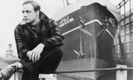 Marlon Brando pictured in black and white, in On the Waterfront, 1954, sitting on railings in front of a large ship.