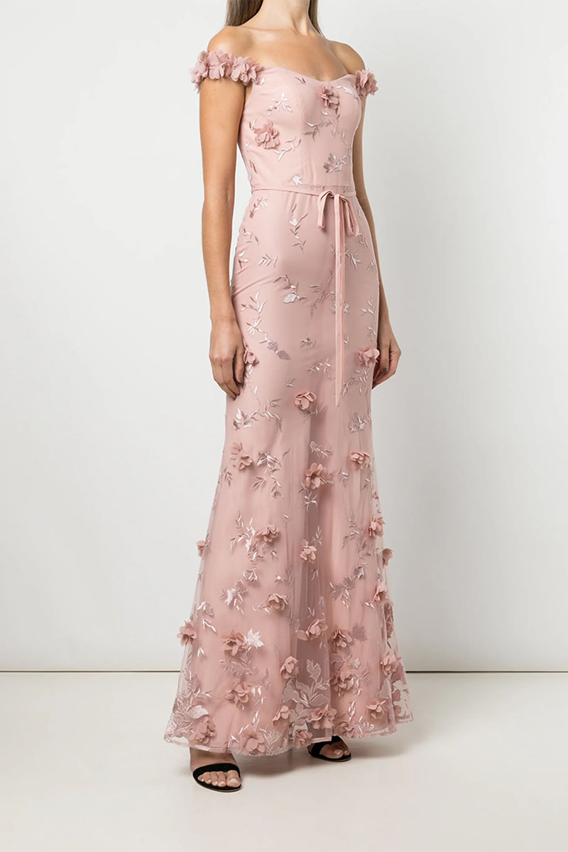 Model wears long pink gown with floral embellishments against grey background. 