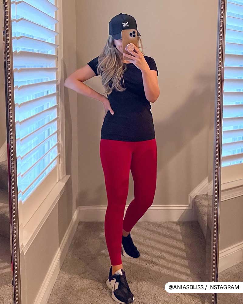 Fashionable woman in black top, red leggings, and cap taking a mirror selfie.