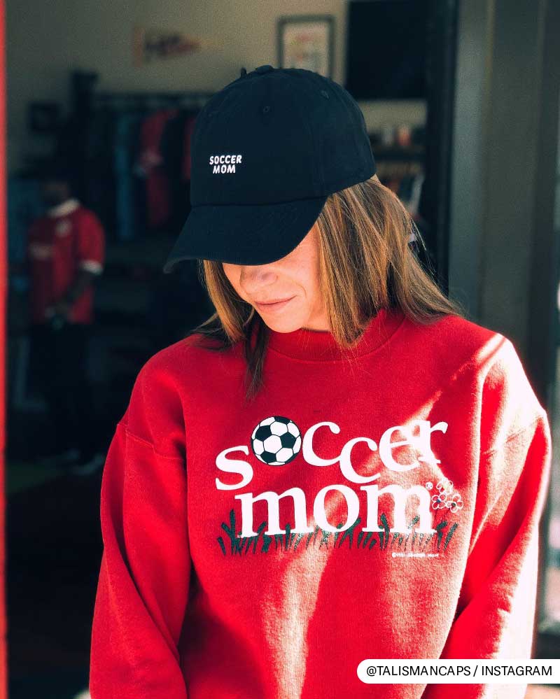 Woman in red 'soccer mom' sweatshirt and black cap, smiling subtly indoors.