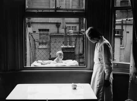 A nanny supervising a baby suspended in a wire cage attached to the outside of a high tenement block window