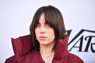 Billie Eilish posing at a Variety event wearing a red coat with a large collar; her brunette hair is styled in a wolf cut