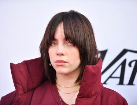 Billie Eilish posing at a Variety event wearing a red coat with a large collar; her brunette hair is styled in a wolf cut