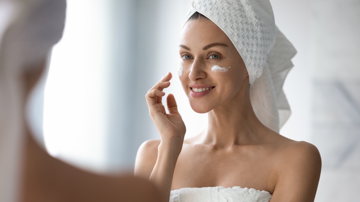 Smiling pretty lady put anti-ageing hydrating moisturizing lifting nourishing facial cream in bathroom, happy young woman wrap in towel look in mirror apply creme on face skin care treatment concept