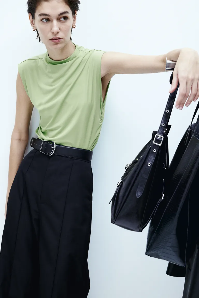 Model wears mint green crop top with black pants and holds handbag. 