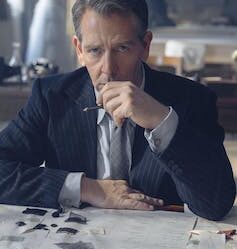 A sombre-looking man sitting at a desk with some fashion designs on the desk in front of him.