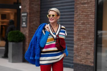 Smiling Happy Model is wearing blue jacket and sweater, striped gilet, red pants and yellow sunglasses. She is walking along the sidewalk.