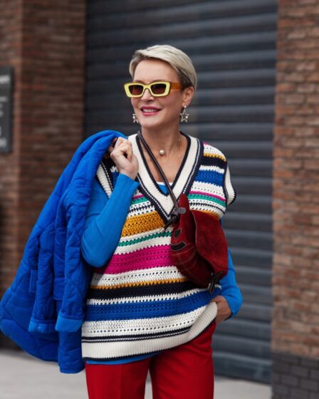 Smiling Happy Model is wearing blue jacket and sweater, striped gilet, red pants and yellow sunglasses. She is walking along the sidewalk.