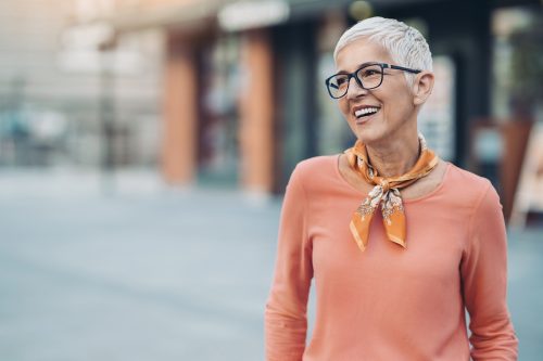 Smiling mature woman with short white hair and eyeglasses wearing a peach shirt while walking outside