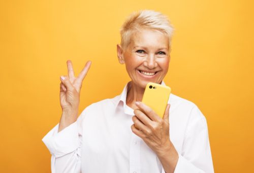 mature woman with a blonde pixie haircut wearing a white blouse giving the peace sign against a yellow background