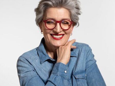 Happy Older Woman with Gray Hair wearing a denim shirt and red glasses against a light gray background