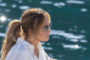 Side profile of Jennifer Lopez wearing a white shirt on the water. Her hair is pulled back into a ponytail and she's wearing sunglasses.