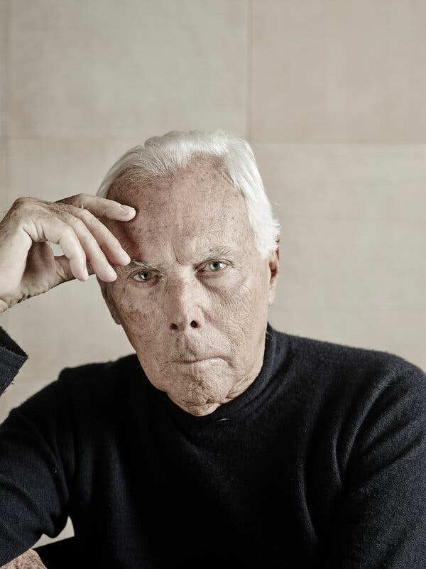 Giorgio Armani stares into the camera with his right hand resting on his forehead. he wears a black sweater and his white hair is combed back. 