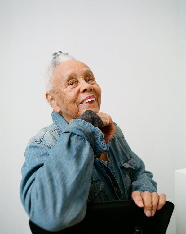 Betye Saar, wearing a denim jacket, looks over her shoulder and smiles. She wears a light red lipstick and her gray hair is tied up on top of her head.
