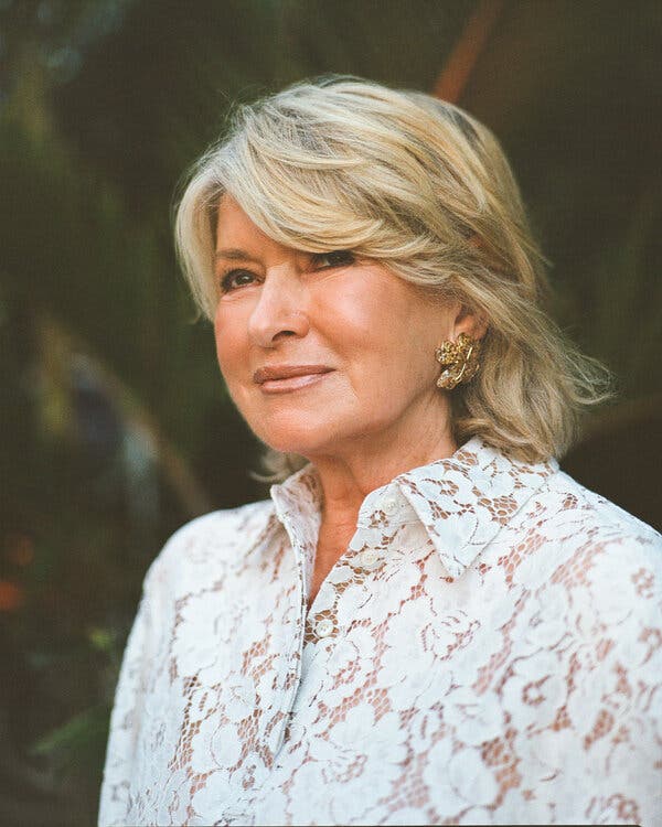 Martha Stewart wears a shirt made of white lace with flower designs and large gold earrings. She looks to the left and her blonde hair swoops just above her eye. 