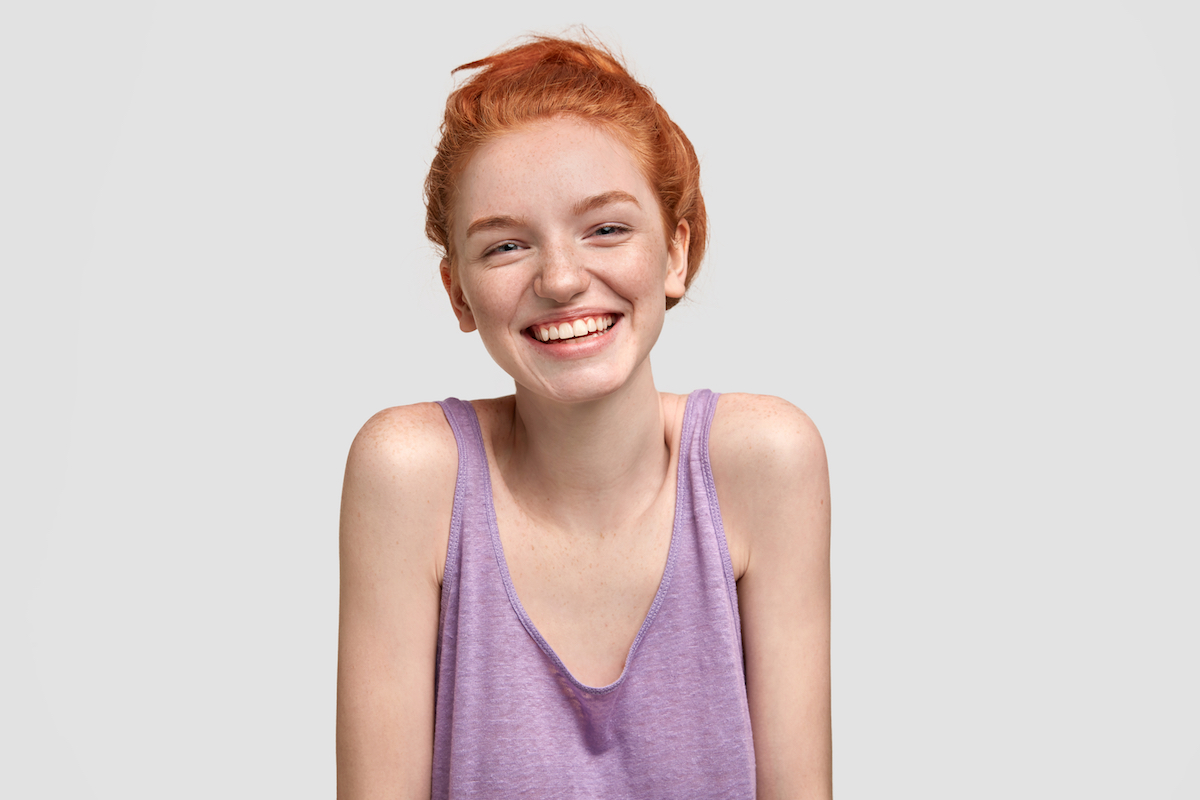 Portrait of happy freckled female with ginger hair, has broad smile, glad to meet with someone, dressed in oversized purple t shirt, isolated over white background. Red haired teenager indoor