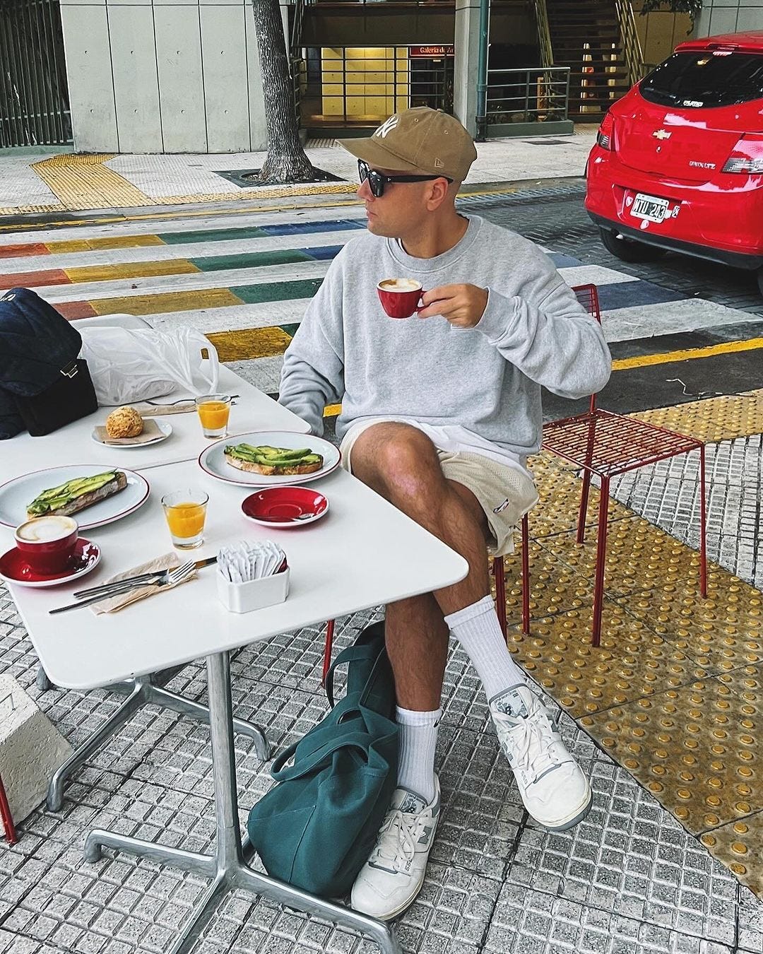 man sitting at an outdoor cafe drinking a coffee and wearing a grey sweatshirt, shorts, and white sneakers with tall socks