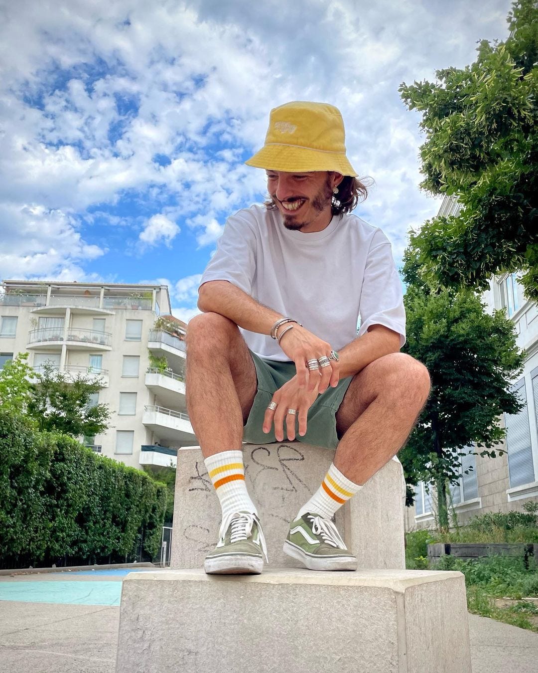 man smiling, wearing a yellow bucket hat, white t-shirt, green shorts, and tall socks with Vans sneakers