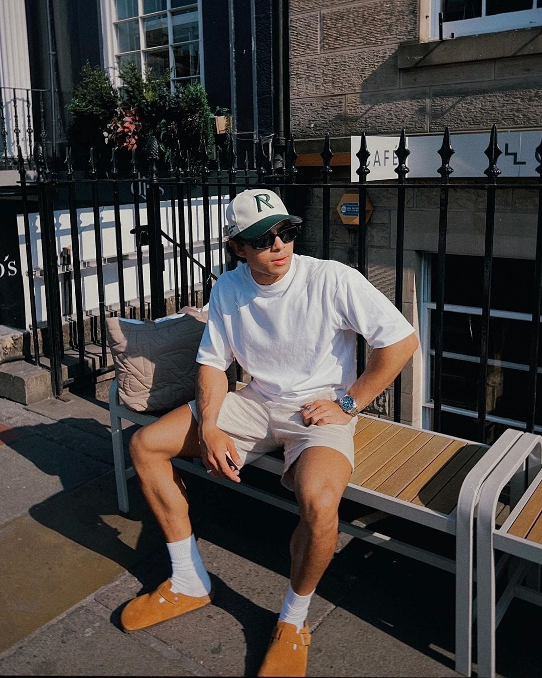 man sitting on a bench outside wearing a baseball hat, white t-shirt, shorts, and tall socks with birkenstock clogs
