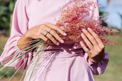 Close shot of romantic cute pink gel polished manicure nails. Woman in pink summer dress holding wild flowers. Two rings on fingers. Outdoor portrait.