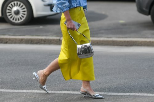 Street style, woman wearing Prada outfit: blue V-neck bra, a matching blue cropped bomber coat, yellow midi skirt and silver pointed pumps heels shoes.